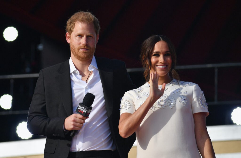 Prince Harry in a black suit on stage with Meghan Markle in a white dress