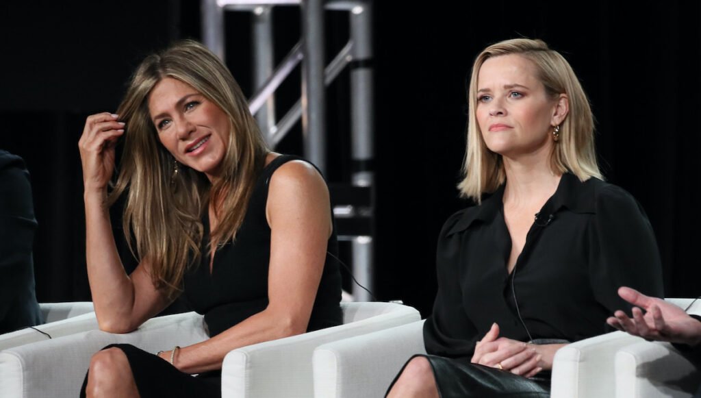 Jennifer Aniston and Reese Witherspoon in black dresses sitting in white chairs