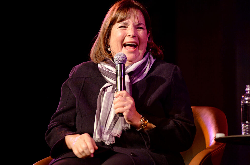 Ina Garten holding a microphone on sage, laughing.