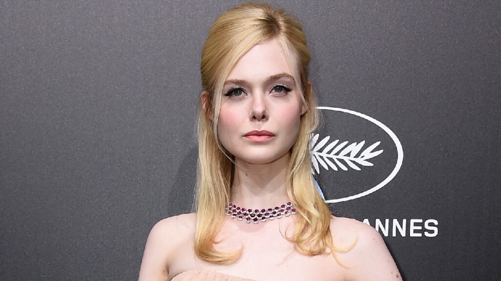 Elle Fanning wears a pale blush colored gown against a gray background on the red carpet