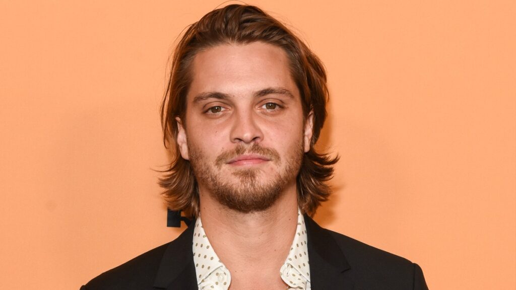 Luke Grimes wears a black suit in front of an orange background on the red carpet