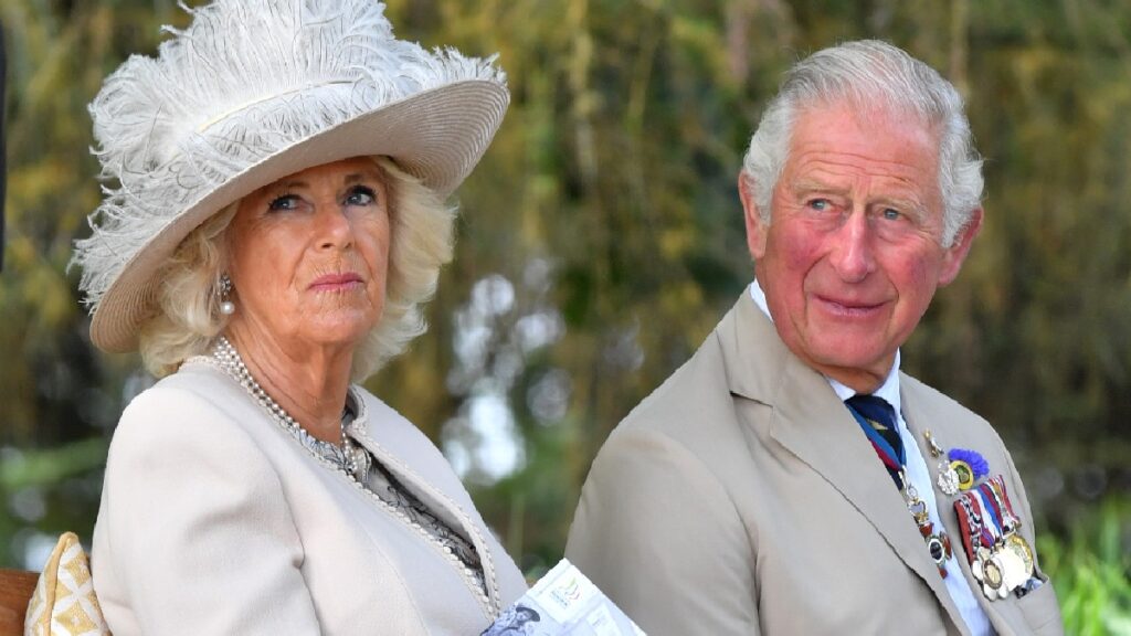 Camilla Parker Bowles and Prince Charles both wear light tan clothes as they sit outdoors on a bench