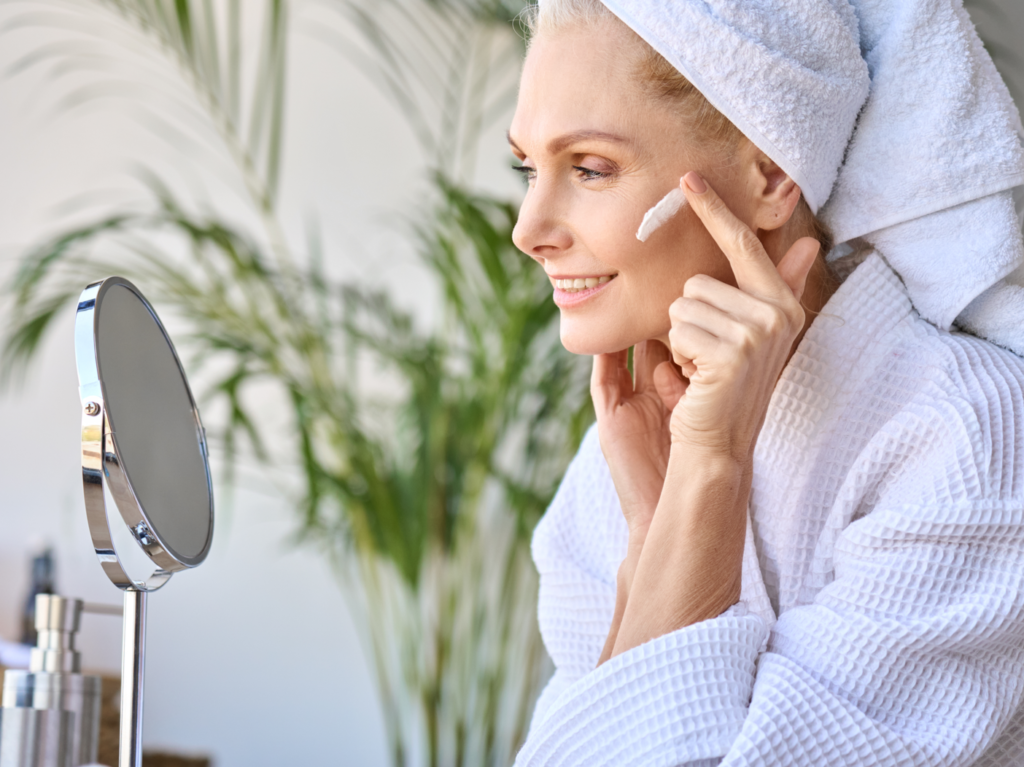 Gorgeous senior older adult 50 years old blonde woman wearing bathrobe and turban towel in bathroom applying moisturizing tightening face skin treatment, looking at mirror. Morning beauty routine.