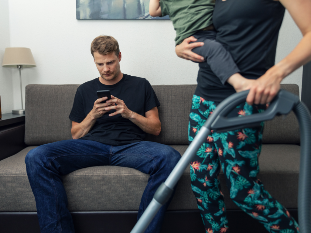 lazy husband sitting in couch and using phone and not helping wife while she cleaning house with child on her lap