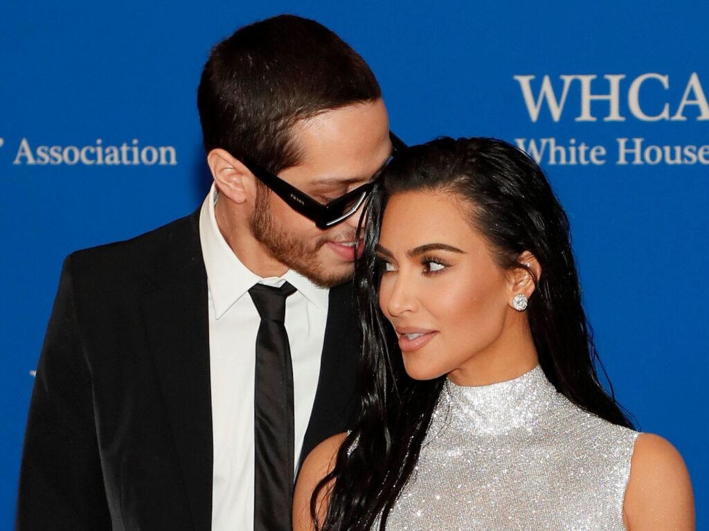 Pete Davidson (L) whispers into Kim Kardashian's ear. He is wearing a black suit and tie and she is wearing a silver mock-neck dress