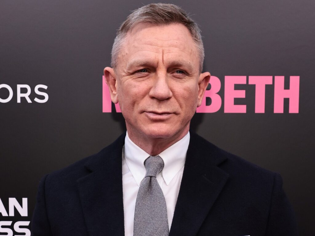 Daniel Craig looks ahead while wearing a white shirt, black blazer, and gray tie
