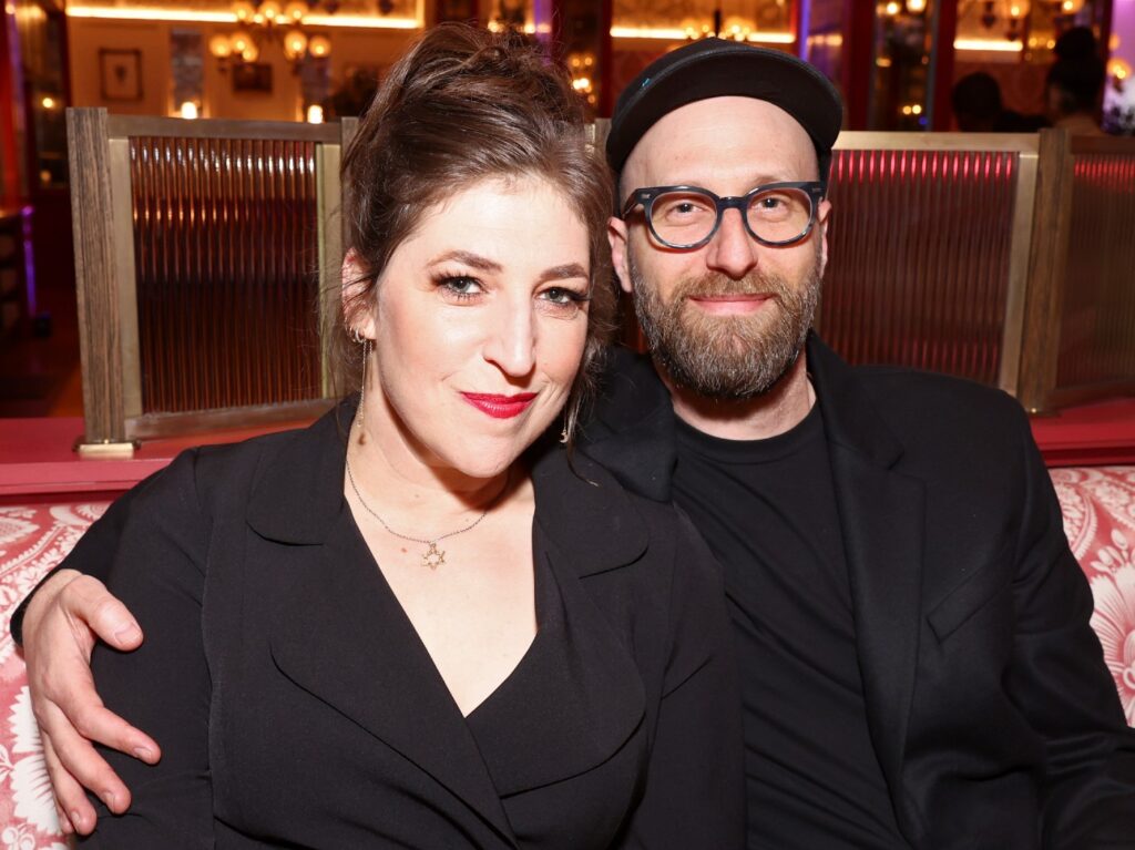 Mayim Bialik (L) and Jonathan Cohen (R) sit at a table. Both are wearing all-black outfits.