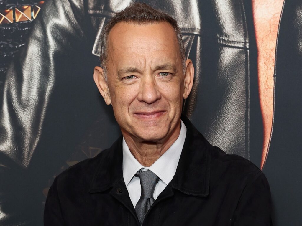 Tom Hanks smiles while wearing a black blazer over a white dress shirt and gray tie