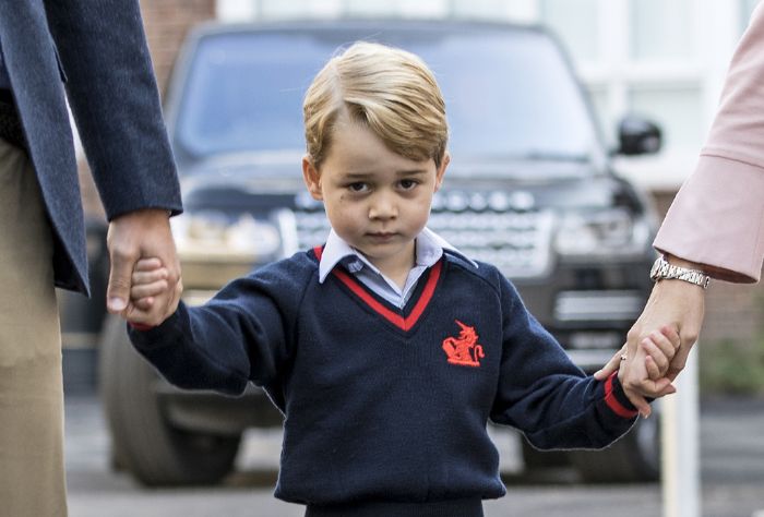 A young boy is looking at the camera while holding two adults' hands. He is wearing a navy sweater with red trim.