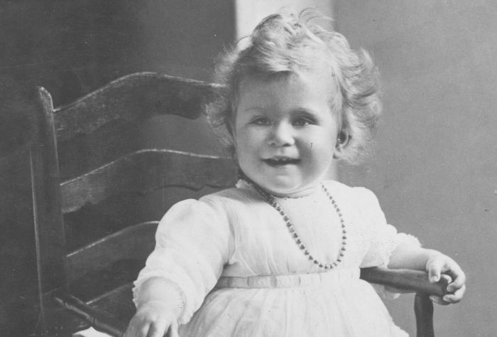 In this black and white photo, a young toddler smiles. She is wearing a light dress with a beaded nacklace.