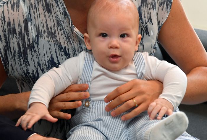 A baby looks at the camera. he is wearing blue gingham overalls over a white long-sleeved shirt