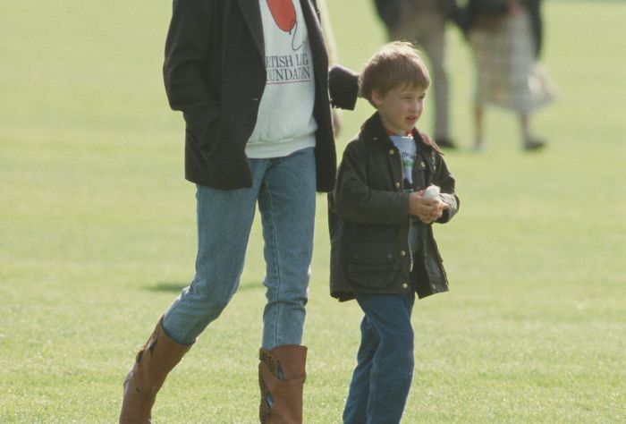 A young boy walks outside with an adult following him, whose face is out of frame. The boy is wearing jeans and a brown jacket