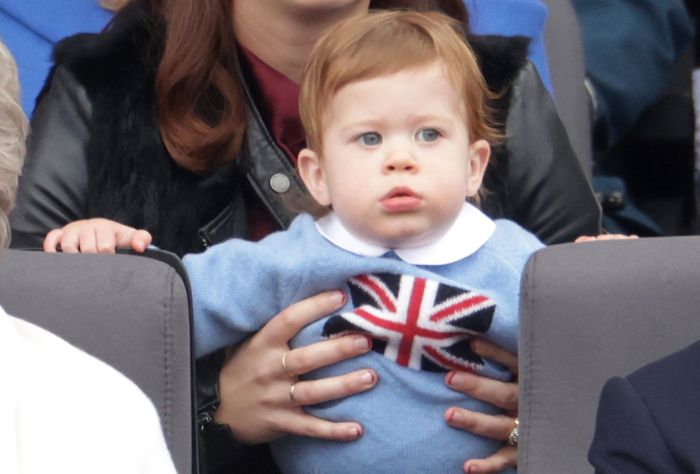 A young red-headed baby looks into the distance. he is wearing a blue sweater with the UK flag on it