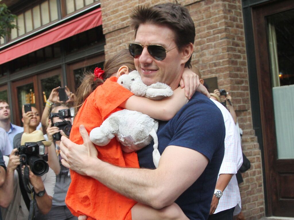 Tom Cruise walks outside in sunglasses and a navy blue shirt. He is carrying his young daughter, whose face is not visible in the photo. She is wearing a bright orange dress