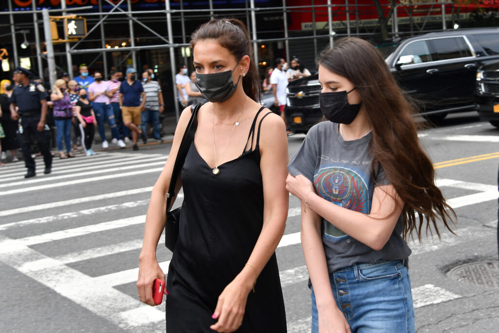 Katie Holmes (L) wearing black tank top and black face mask, crossing a street with Suri Cruise, who is wearing a gray graphic tee with jeans and a black face mask.