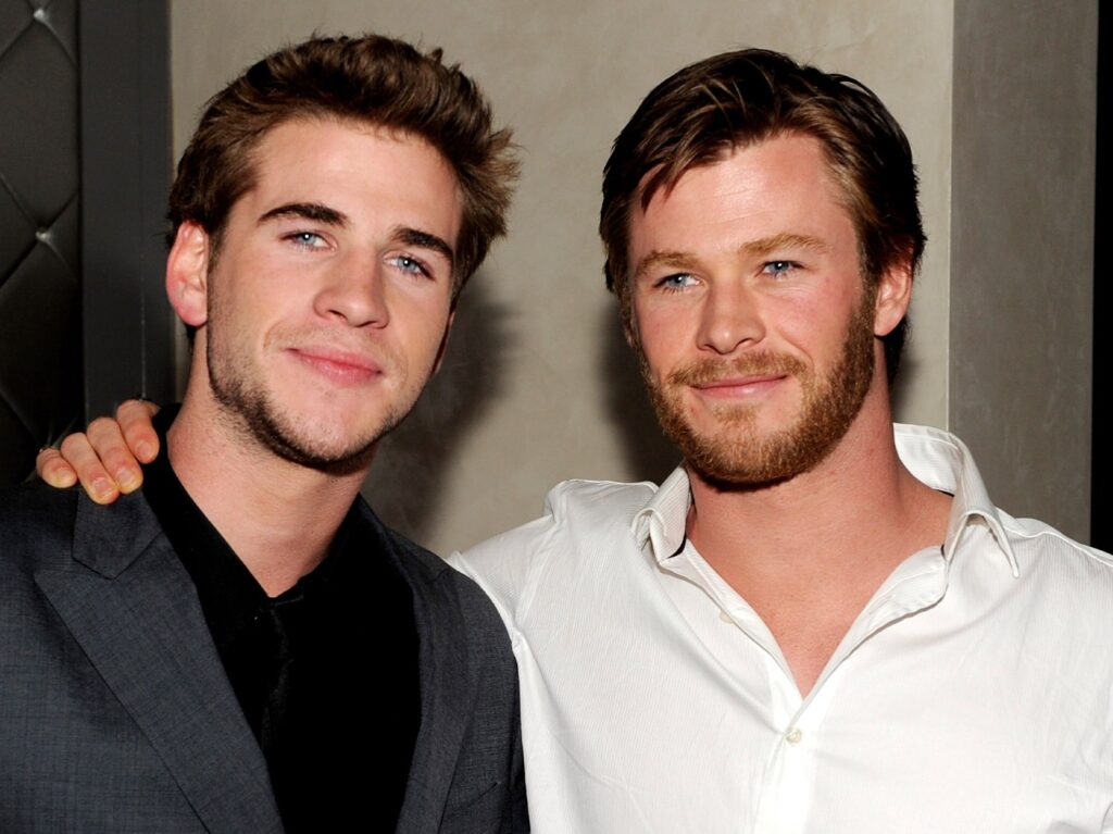 Liam Hemsworth (LO wearing black top and gray blazer, standing next to Chris Hemsworth, who is wearing a white dress shirt