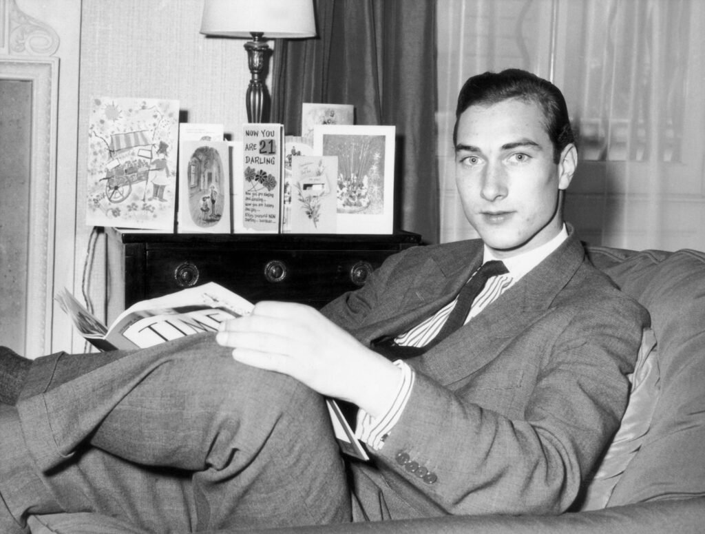 Black and white image of Prince William of Gloucester sitting on a couch and holding a book in his lap. He is dressed in a suit and tie.