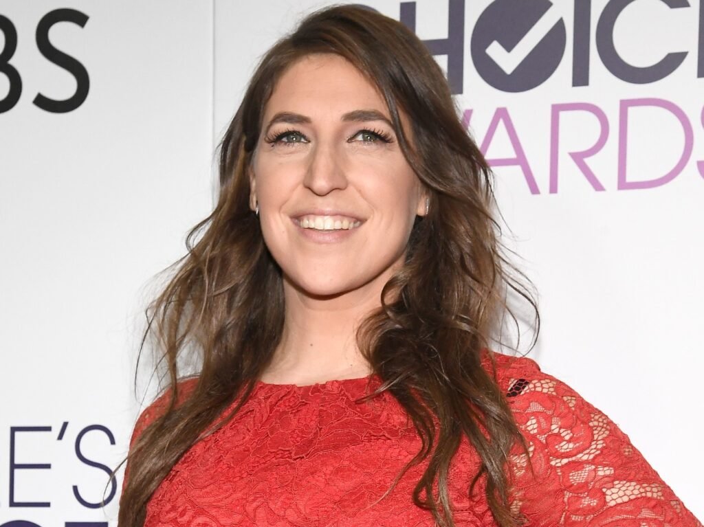 Mayim Bialik smiles while wearing red longsleeved lace dress