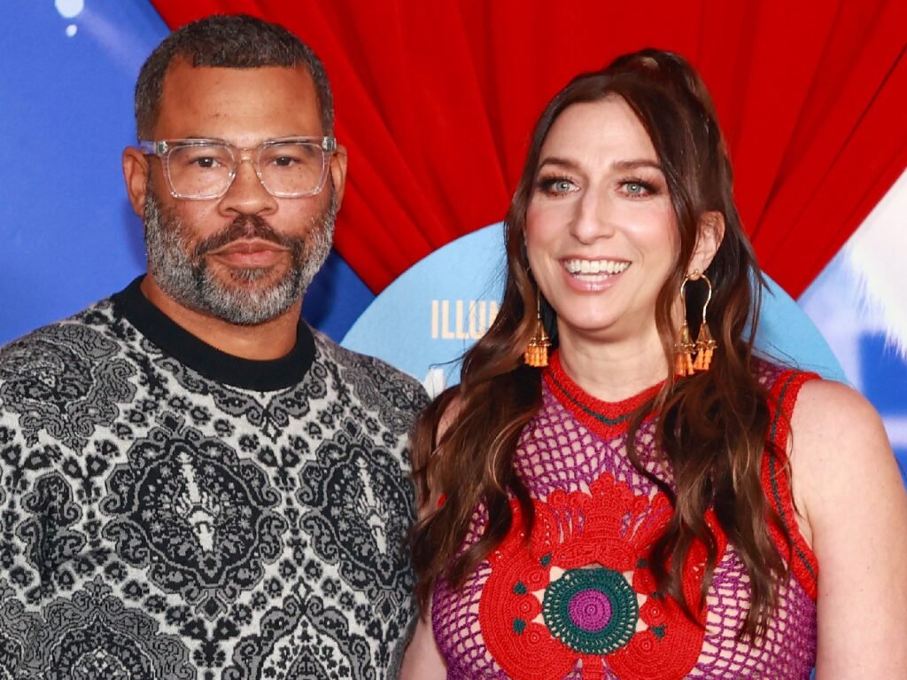 Jordan Peele (L) wearing gray patterned sweater, standing next to a smiling Chelsea Peretti, who is wearing a red patterned top