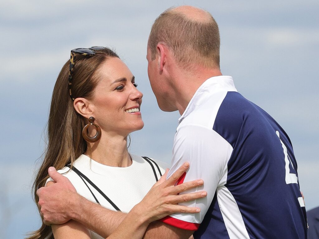 Kate Middleton (L) smiles and embraces Prince William. Middleton is wearing a white dress with black pinstripes and William is wearing a jersey