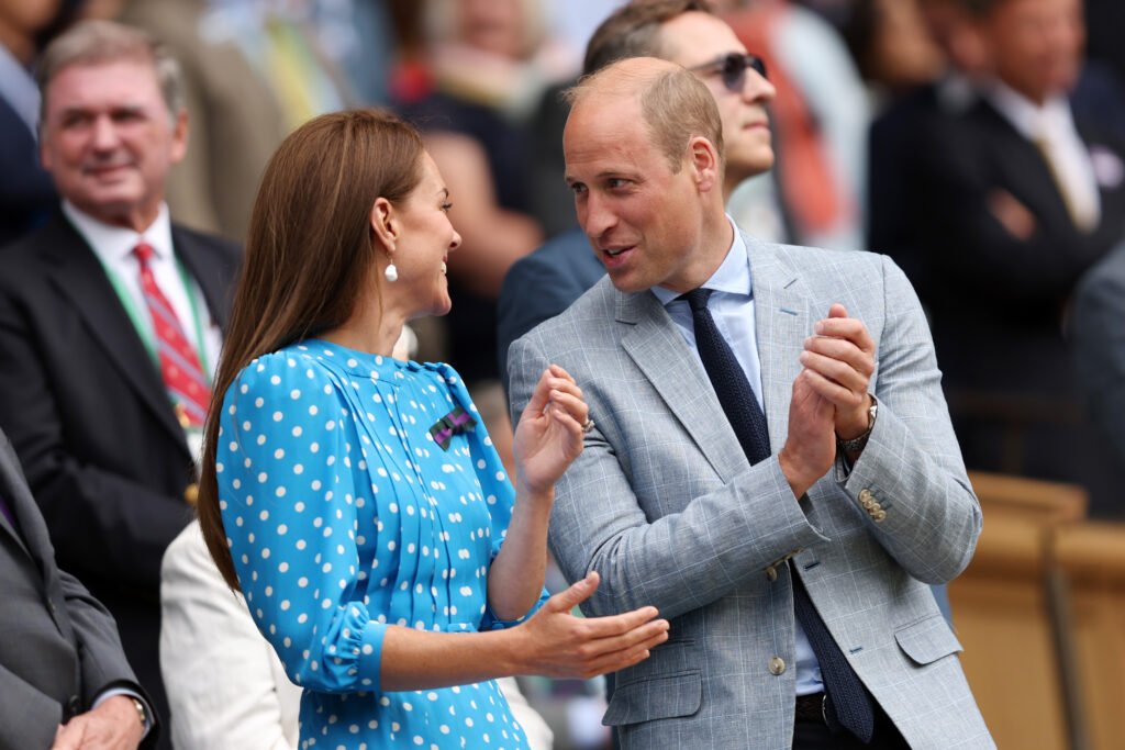 Kate Middleton (L) laughs while looking at Prince William. Middleton is wearing a bright blue dress with white polka dots, while William is wearing a gray blazer over a light dress top and black tie