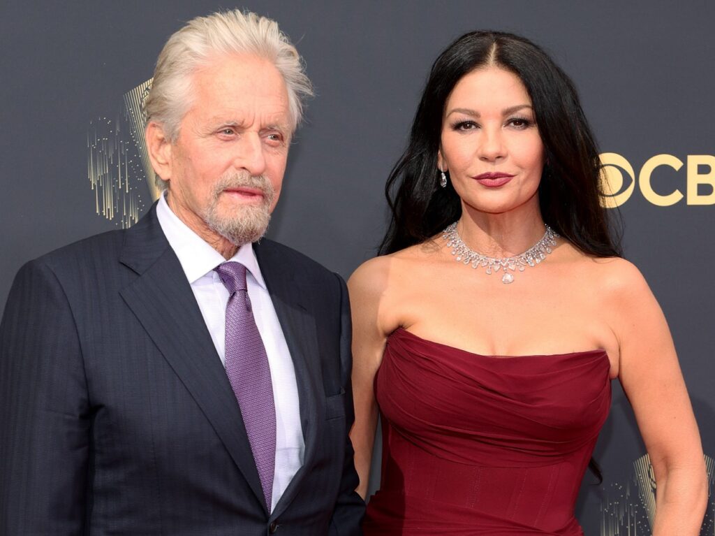Michael Douglas (L) wearing black blazer over white dress shirt and purple tie, standing next to Catherine Zeta-Jones, who is wearing a deep red gown