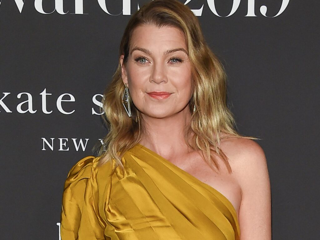 Ellen Pompeo smirks while wearing one-shoulder gold jumpsuit against charcoal gray background