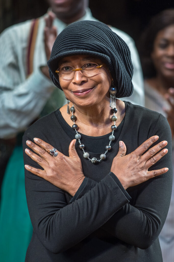 Alice Walker smiling with her hands crossed over her chest in a black outfit with matching hats t