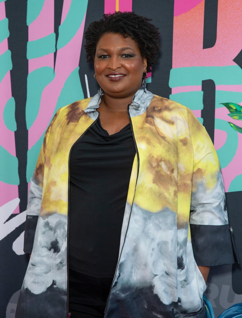 Stacey Abrams smiling in black shirt with yellow jacket