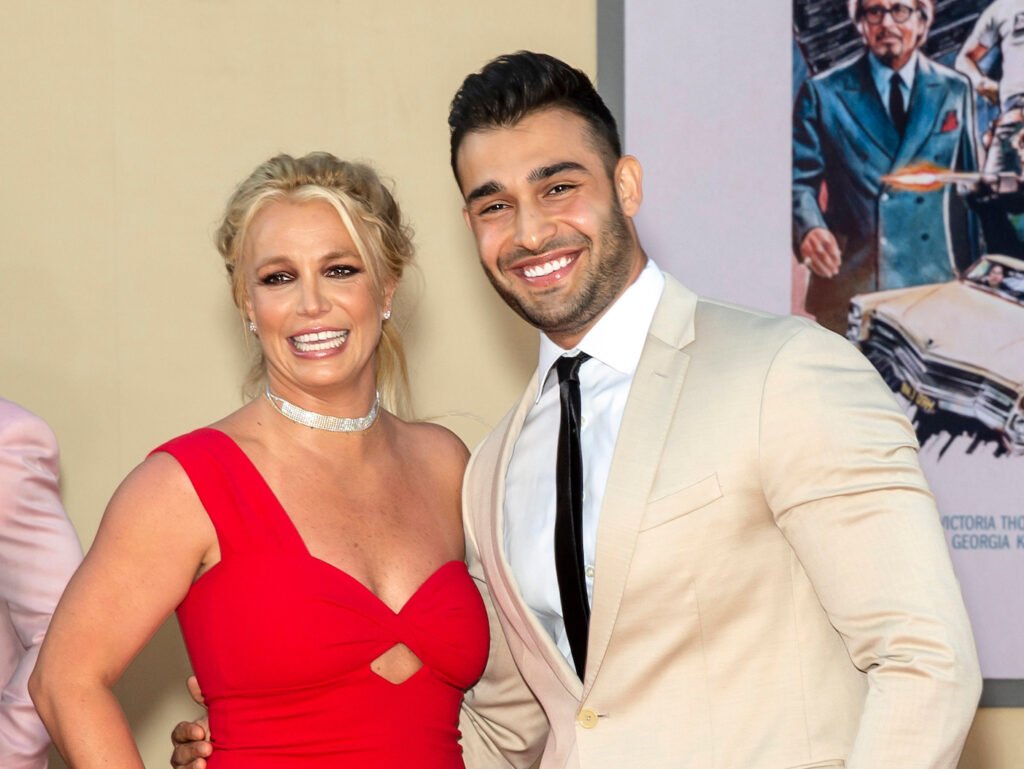 2019 photo of Britney Spears smiling in a red dress with Sam Asghari in a tan suit