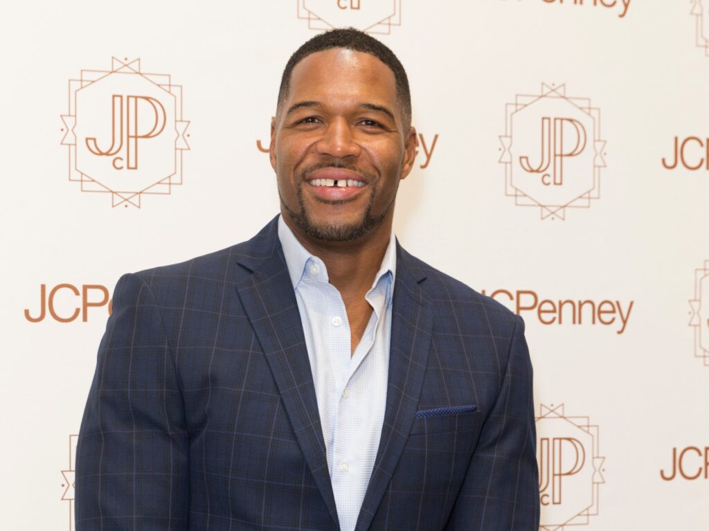 Michael Strahan smiling in blazer and dress shirt against cream backdrop