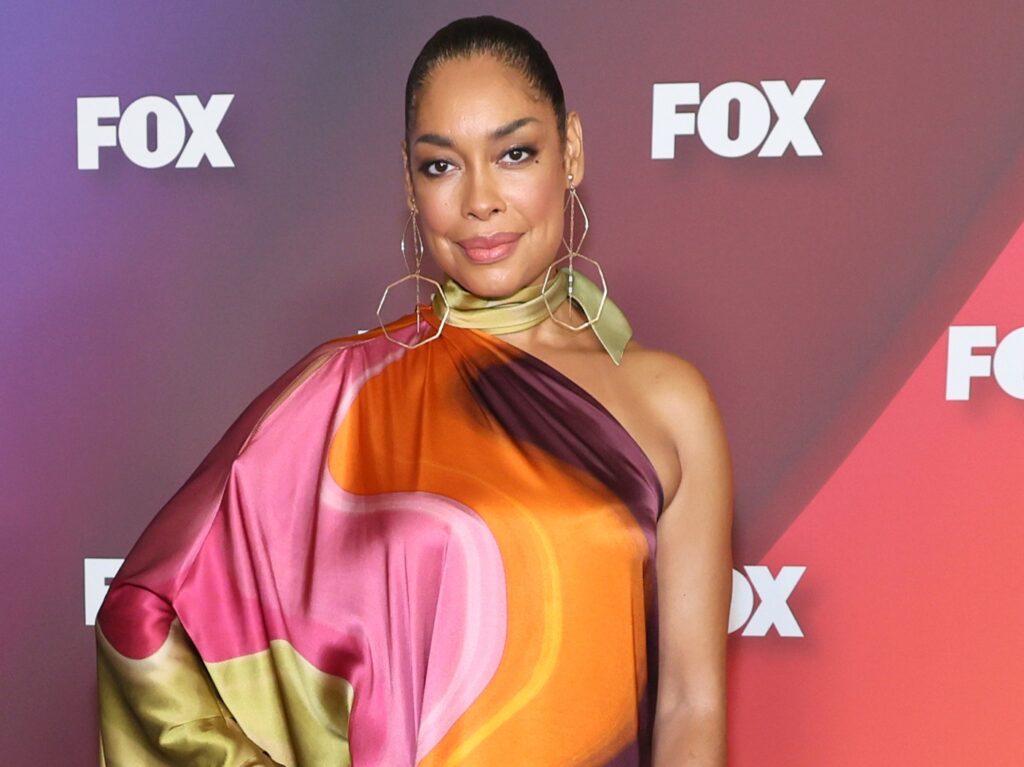 Gina Torres smiles in multi-patterned dress against purple backdrop