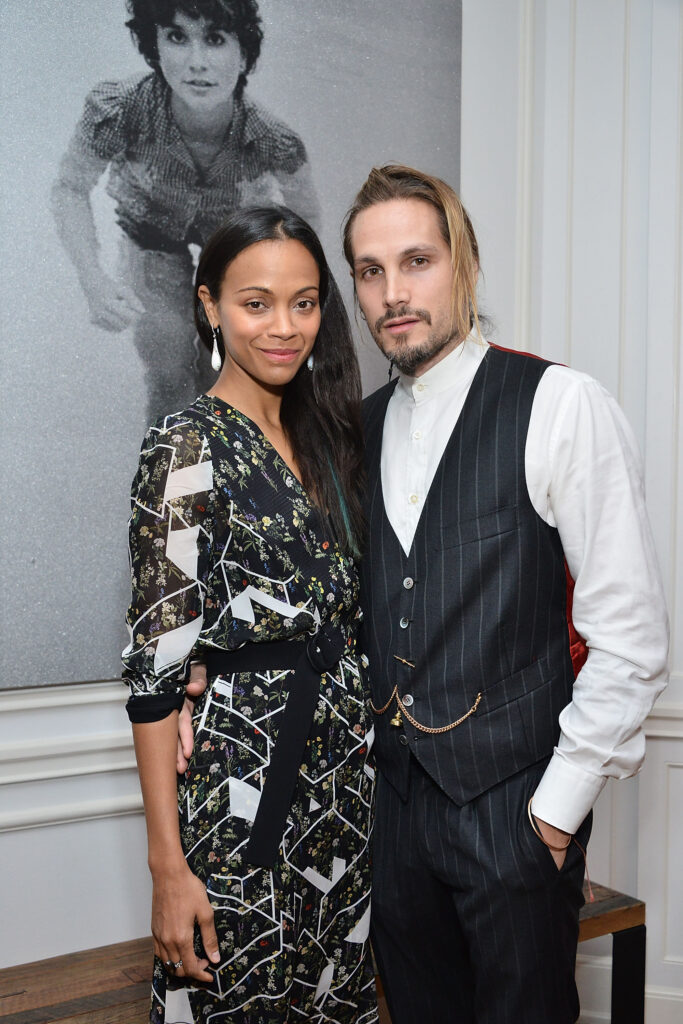 Mr. and Mrs. Saldana attend an art exhibition. Zoe wears a patterned black and white dress and Marco wears a striped tuxedo over a white dress shirt