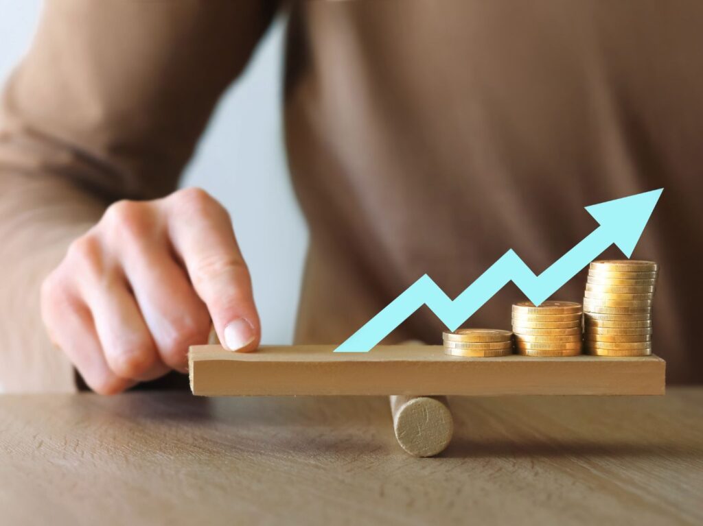 Person holds finger down on balance, stack of coins on the other side with blue arrow on top pointing upward to signify inflation