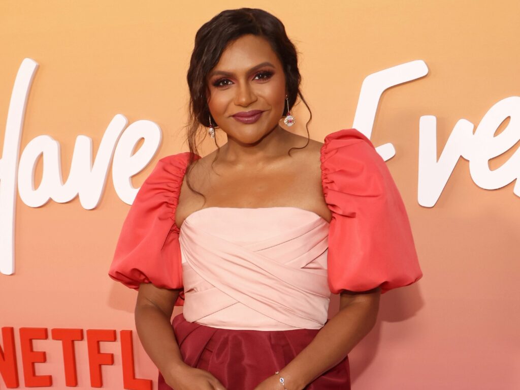 Mindy Kaling smiles in multi-colored dress against orange backdrop