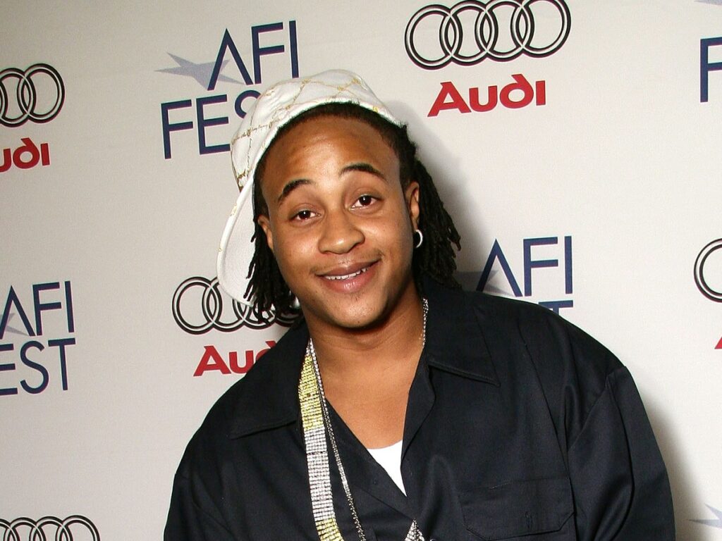 Orlando Brown smiles in black jacket and white baseball cap against white backdrop