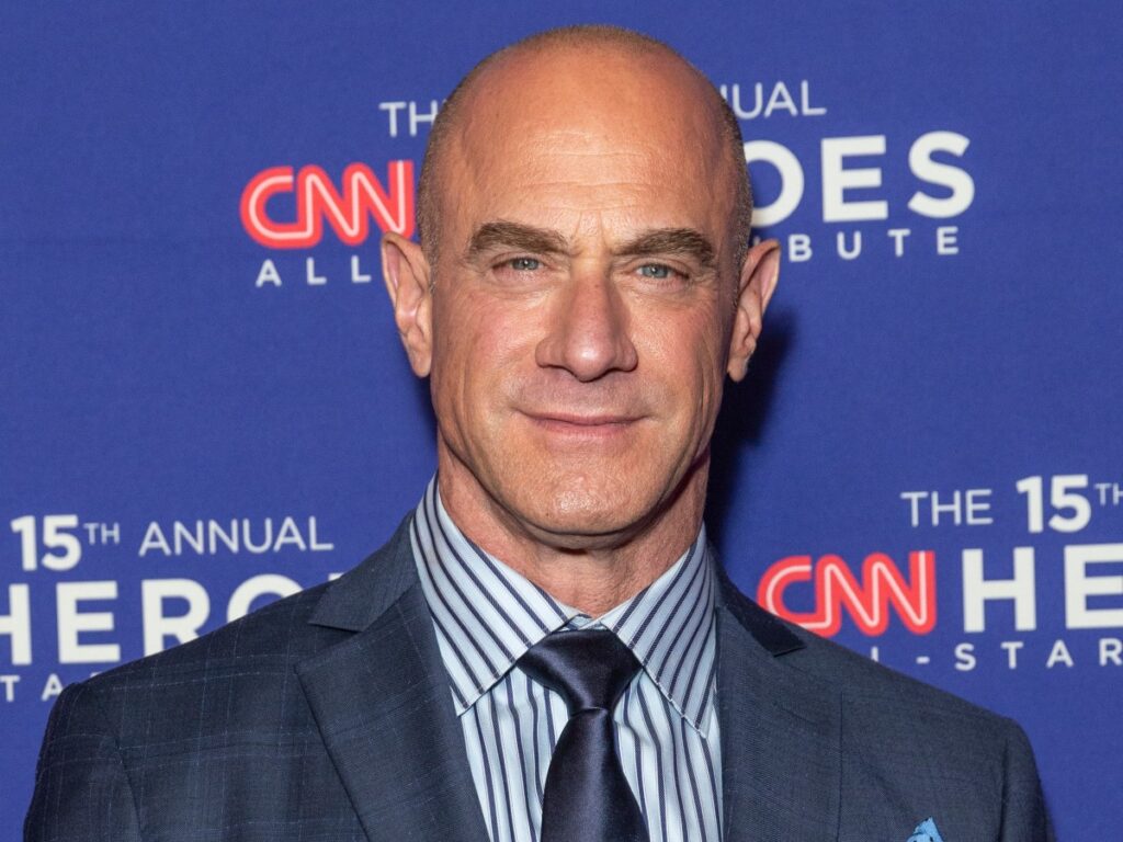 Christopher Meloni smiles in dark blue suit with striped shirt and tie against blue backdrop