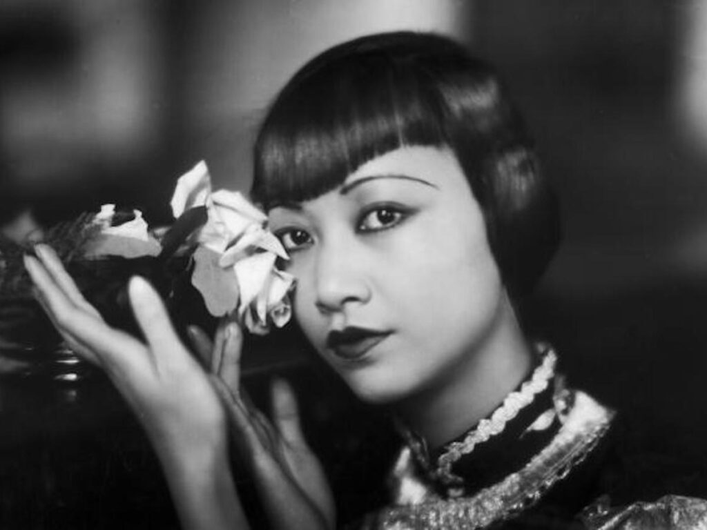 1935 black and white photo of Anna May Wong with bangs and gesturing to a rose