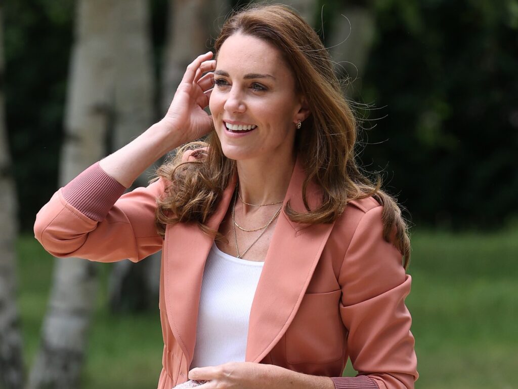 Kate Middleton smiles and pushes her hair behind her ear. She is wearing a white top and salmon-colored jacket