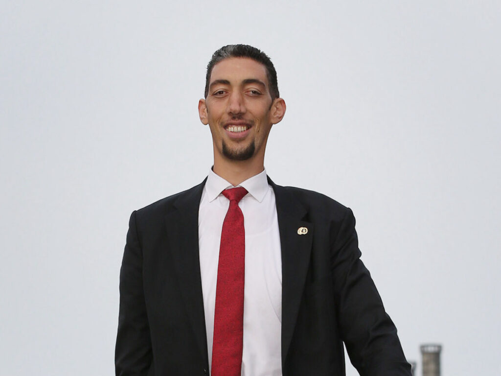 Sultan Kosen smiling brightly in a black suit with white tie outdoors on an overcast day