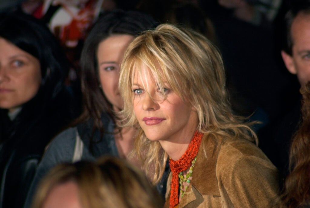 Meg Ryan in a crowd of people wearing a brown jacket and red scarf.