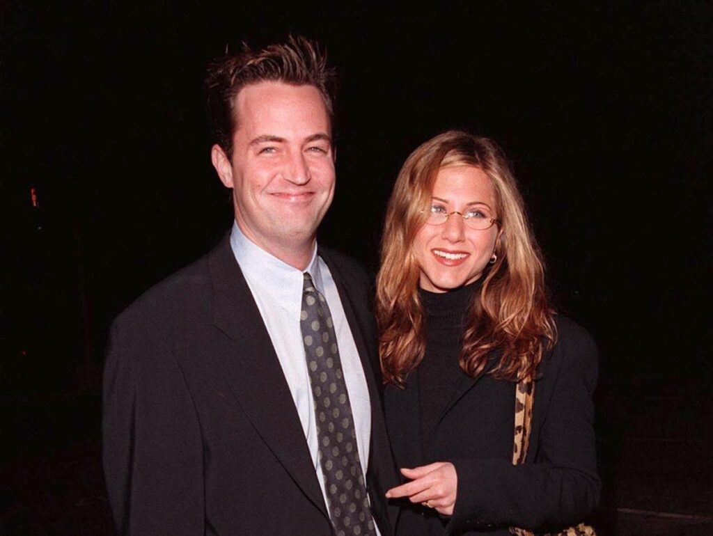 1998 photo of Matthew Perry in a large brown suit and Jennifer Aniston in a black turtleneck and wire glasses
